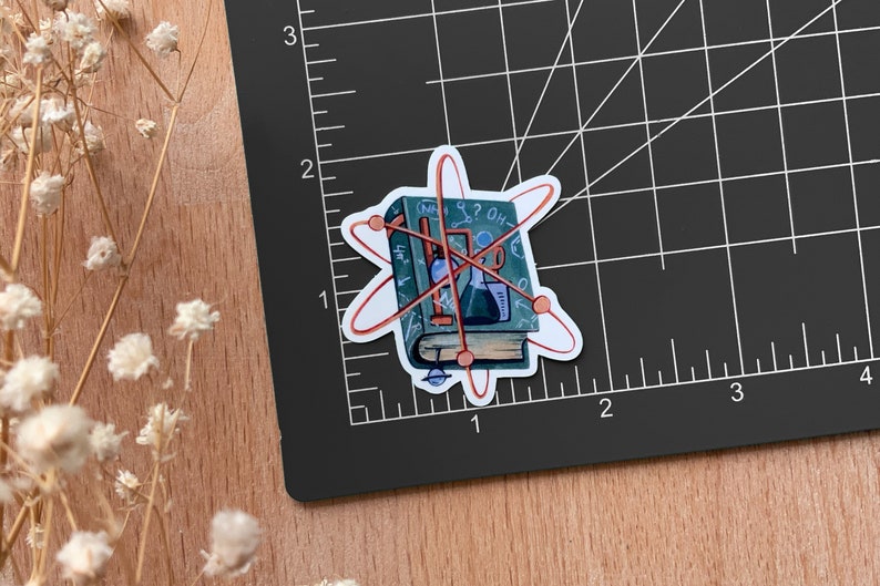 May include: A sticker of a closed book with a red atomic symbol around it. The book has a blue cover with handwritten notes and drawings of scientific equipment, including a beaker, a flask, and a graduated cylinder. The book is titled "The Book of Science".