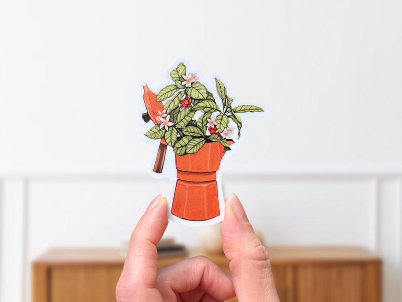 Peut inclure: Un autocollant repr&eacute;sentant une cafeti&egrave;re orange avec une plante qui en sort. La plante a des feuilles vertes, des fleurs blanches et des baies rouges. L'autocollant est tenu entre deux doigts sur un fond blanc.