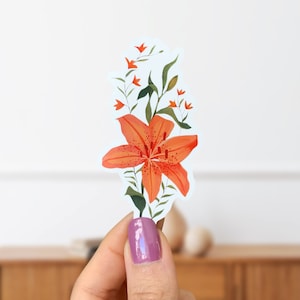 Puede incluir: Una pegatina con una ilustración de acuarela de un lirio naranja con hojas y tallos verdes. El lirio está en flor y tiene un centro naranja prominente.