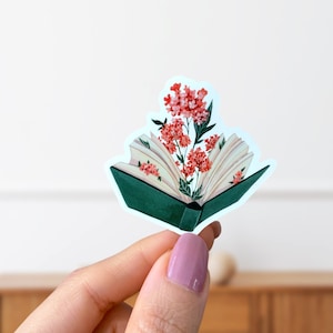 May include: A green book with its pages open, revealing a bouquet of pink flowers. The book is a sticker with a white outline.