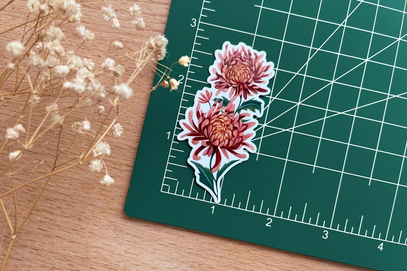 Peut inclure: Un autocollant repr&eacute;sentant un motif floral avec deux chrysanth&egrave;mes roses et des feuilles vertes sur fond blanc.