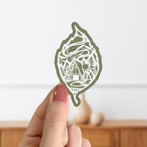 May include: A green leaf-shaped sticker featuring a white illustration of a cabin in the woods. The cabin has a triangular roof and windows. There are trees and a winding road leading to the cabin. Smoke is rising from the chimney.