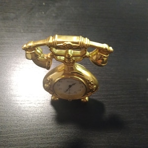 May include: Gold-toned desk clock shaped like a vintage rotary phone. The clock face has black hands and black Roman numeral markings.