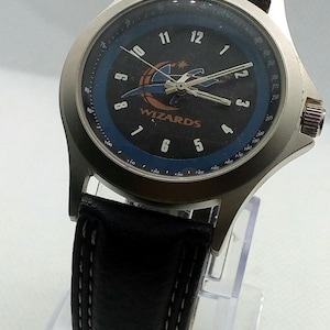 May include: A silver watch with a black face and a black leather band. The watch face has the Washington Wizards logo and the words "WIZARDS" printed on it. The watch has a tachymeter scale around the outer edge of the face.