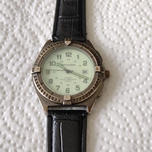 May include: A silver-toned wristwatch with a black leather strap. The watch face is white with black numbers and hands. The watch is water resistant and has a quartz movement. The watch face also says "MILAN" and "WATER RESISTANT QUARTZ".