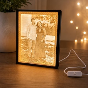 Pode incluir: Uma moldura de foto iluminada com borda preta exibe uma imagem em tons sépia de um casal. A moldura está conectada com um cabo branco e possui um interruptor liga/desliga. A moldura está sobre uma superfície de madeira.