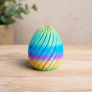 Könnte beinhalten: Ein mehrfarbiges, segmentiertes Osterei in Grün-, Blau-, Lila- und Gelbtönen. Das Ei hat eine strukturierte, geriffelte Oberfläche mit einem dekorativen Muster. Das Ei steht auf einer hellen Holzoberfläche.