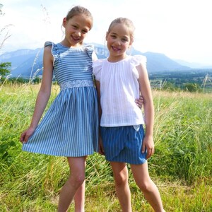 Peut inclure: Deux filles portant des robes à rayures bleues et blanches se tiennent dans un champ d'herbe verte. La fille à gauche porte une robe à rayures bleues et blanches avec une bordure en dentelle blanche. La fille à droite porte un haut blanc avec une jupe à rayures bleues et blanches.
