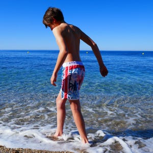 May include: A young person wearing colorful swim trunks with a tropical print walks through the shallow water of a beach.