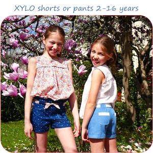 Peut inclure: Deux filles portant des shorts bleus avec des bordures blanches et des hauts à imprimé floral. Les filles sont debout devant un arbre avec des fleurs roses. Le texte "XYLO shorts ou pantalons 2-16 ans" est en haut de l'image.
