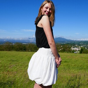 Könnte beinhalten: Eine junge Person trägt ein schwarzes ärmelloses Oberteil und einen weißen Ballonrock. Die Person steht auf einer grasbewachsenen Wiese mit einem Gebirge im Hintergrund.