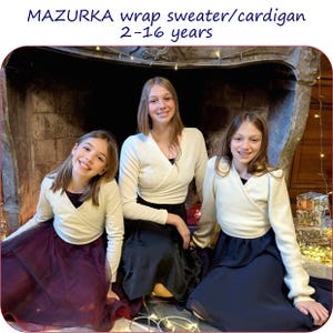 Puede incluir: Tres personas visten suéteres cruzados color crema y faldas oscuras frente a una chimenea. Los suéteres tienen un diseño cruzado. El texto "MAZURKA wrap sweater/cardigan 2-16 years" está en la parte superior.