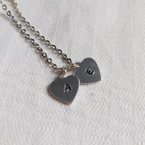 May include: A silver necklace featuring two heart-shaped pendants. Each pendant has a stamped initial, with the letters "A" and "E" visible. The chain is a delicate link style, and the pendants are suspended from it.