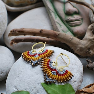 May include: A pair of gold hoop earrings with a beaded design. The beads are red, yellow, and grey. The earrings are on a white surface.