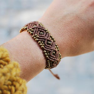 May include: A brown and gold macrame bracelet with a geometric pattern. The bracelet is tied with a knot and has a tassel.