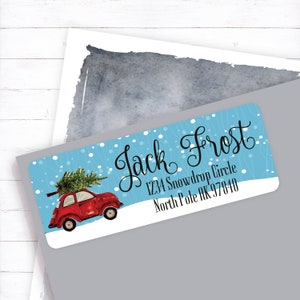May include: Red vintage car with a Christmas tree on top driving through a snowy winter scene. The car is on a blue background with white snowflakes. The text "Jack Frost" is written in cursive above the car. The address "1234 Snowdrop Circle, North Pole AK 97840" is written below the car.