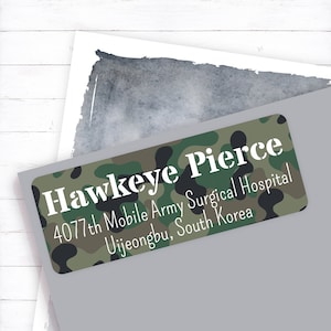 May include: A rectangular sticker with a camouflage pattern in shades of green, brown, and black. The sticker features the name "Hawkeye Pierce" in white, along with address details: "4077th Mobile Army Surgical Hospital, Uijeongbu, South Korea."