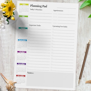 May include: A white planning pad with a calendar for June through December, with sections for priorities, tasks, and notes. A brown pen rests on the right side. The pad is on a light wood-grain surface.