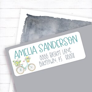 May include: A white return address label with a teal bicycle carrying a basket of flowers. The text on the label reads "Amelia Sanderson, 8066 Basket Lane, Biketown, KS 96874."