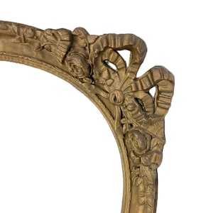 May include: An oval-shaped picture frame with a metallic gold finish. The frame features ornate, raised relief carvings of roses and flowing ribbon details along the outer edge, creating a textured and dimensional appearance on the surface.
