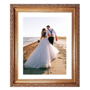 May include: Ornate gold-colored picture frame with a detailed, scrolling design. The frame surrounds a photograph of a couple walking on a beach. The couple is embracing, with the bride in a white wedding dress.