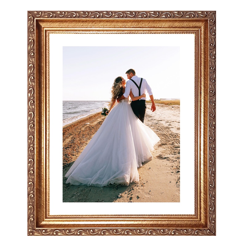 May include: Ornate gold-colored picture frame with a detailed, scrolling design. The frame surrounds a photograph of a couple walking on a beach. The couple is embracing, with the bride in a white wedding dress.