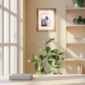 May include: A framed photograph of a wedding couple walking on a beach. The frame is gold with ornate detailing and a white mat.