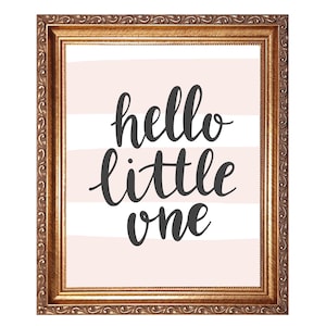 May include: A framed print with the words "hello little one" in dark gray script on a light pink and white striped background. The frame is ornate with a gold finish, adding a touch of elegance to the nursery decor.