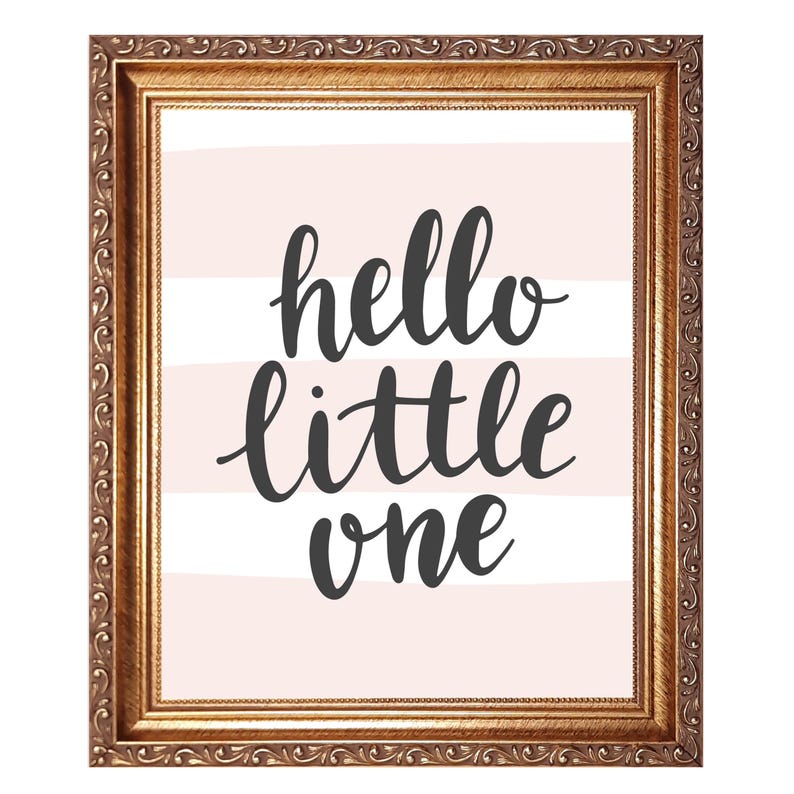 May include: A framed print with the words "hello little one" in dark gray script on a light pink and white striped background. The frame is ornate with a gold finish, adding a touch of elegance to the nursery decor.