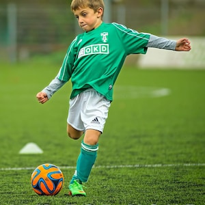 May include: A young soccer player in a green jersey with white shorts and teal socks kicks a soccer ball on a green field. The ball is orange and blue. Text at the top reads "LIAM 2025" and at the bottom "AIM FOR YOUR GOALS."