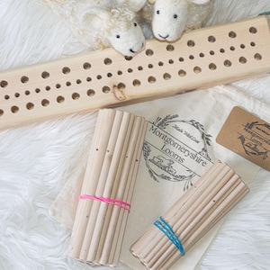 May include: A wooden weaving loom with multiple holes, two bundles of wooden dowels secured with pink and blue elastic bands, and a tag with the text "Montgomeryshire Looms". The items are on a white, fluffy surface.
