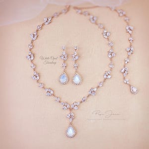 Op de afbeelding: Een roségouden sieradenset bestaande uit een ketting en bijpassende oorbellen met witte opalen druppelstenen en heldere marquise-geslepen kristallen in een bladvormig patroon. De tekst White Opal Teardrop en Pipa Joux Personalized Jewelry Collection is zichtbaar.