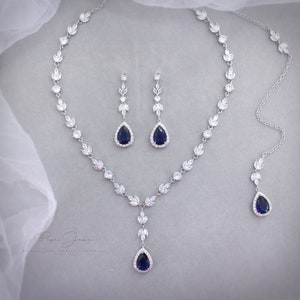 以下が含まれることがあります： 青いサファイアをセットした涙型のペンダントが付いたシルバーネックレス。ネックレスは小さなクリアストーンで飾られており、繊細な葉のデザインが施されています。青いサファイアとクリアストーンをセットしたマッチングイヤリングも含まれています。
