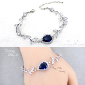 May include: Silver teardrop bracelet with a navy blue teardrop-shaped stone surrounded by smaller clear stones. The bracelet features a delicate vine design with leaves and small clear stones.