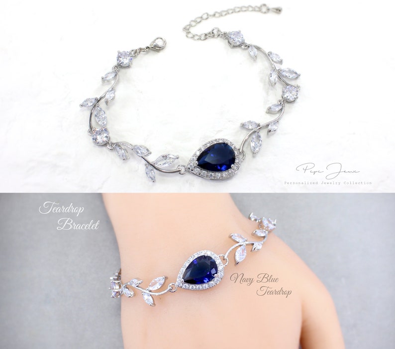 May include: Silver teardrop bracelet with a navy blue teardrop-shaped stone surrounded by smaller clear stones. The bracelet features a delicate vine design with leaves and small clear stones.