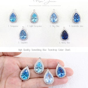 May include: Color chart of teardrop-shaped crystal pendants in various shades of blue, including turquoise, light turquoise, sky blue, sapphire, and navy blue. The pendants are set in silver and have a halo of small clear crystals around the center stone. The text "High Quality Something Blue Teardrop Color Chart" is printed below the pendants.