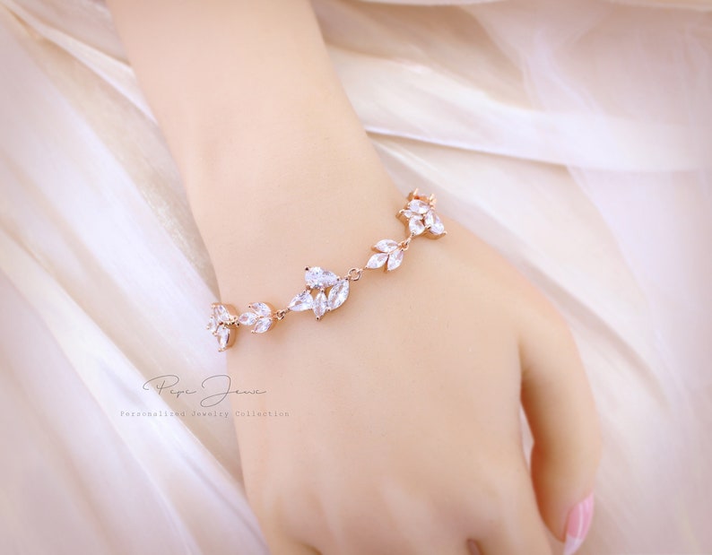 Puede incluir: Un brazalete de oro rosa con un dise&ntilde;o delicado que presenta piedras de circonita c&uacute;bica transparentes dispuestas en forma de hoja. El brazalete se muestra en la mu&ntilde;eca de una modelo.