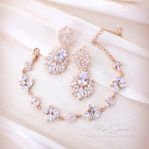 Puede incluir: Un conjunto de joyas de novia de oro rosa con un brazalete delicado y un par de pendientes colgantes. Las joyas presentan piedras de circonita c&uacute;bica transparentes en un dise&ntilde;o floral. El conjunto se muestra sobre un fondo de tela blanca.  Personalized Jewelry Collection.