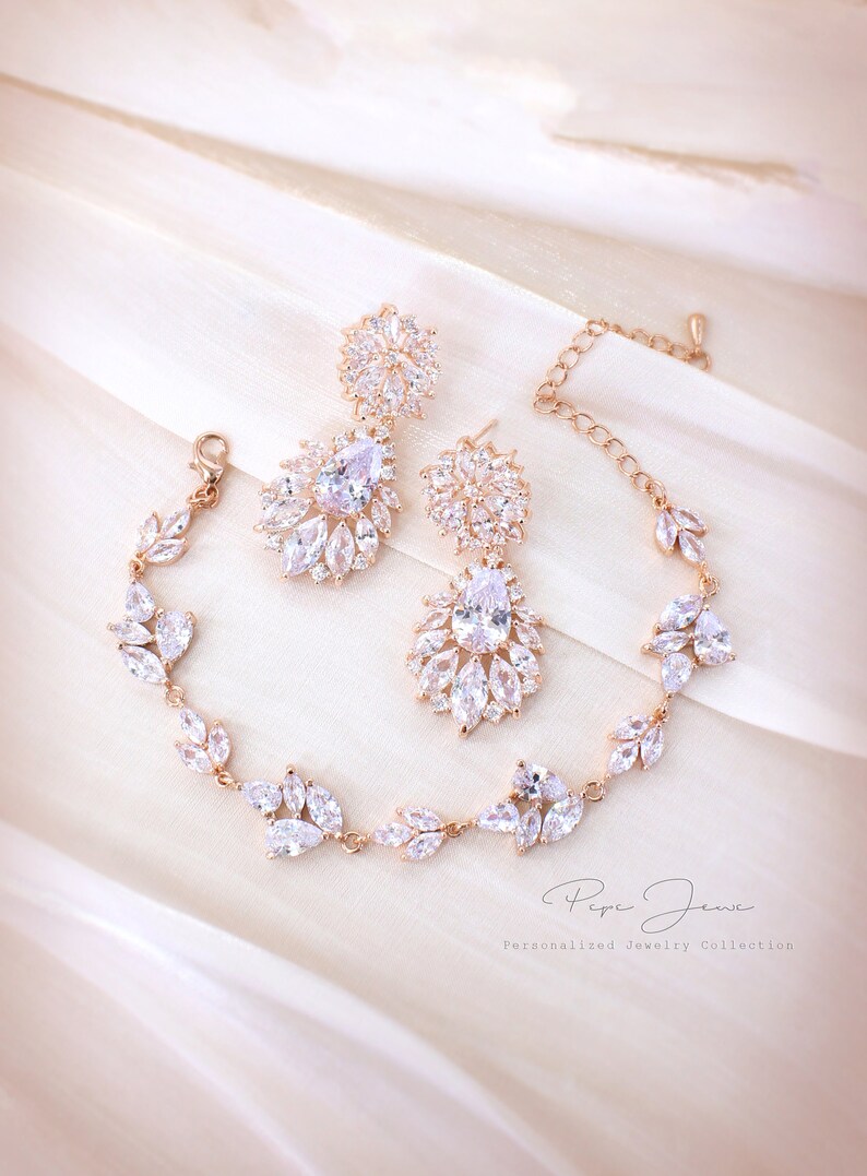 Puede incluir: Un conjunto de joyas de novia de oro rosa con un brazalete delicado y un par de pendientes colgantes. Las joyas presentan piedras de circonita c&uacute;bica transparentes en un dise&ntilde;o floral. El conjunto se muestra sobre un fondo de tela blanca.  Personalized Jewelry Collection.