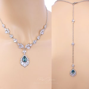 Puede incluir: Un collar de plata con un colgante de piedra preciosa azul en forma de l&aacute;grima rodeado de piedras transparentes m&aacute;s peque&ntilde;as. El collar tiene una cadena delicada con peque&ntilde;as piedras transparentes en forma de hojas. El collar se muestra en dos vistas: la delantera y la trasera. La vista trasera muestra el collar extendi&eacute;ndose hacia abajo por la espalda con una cadena m&aacute;s larga y una versi&oacute;n m&aacute;s peque&ntilde;a del colgante.