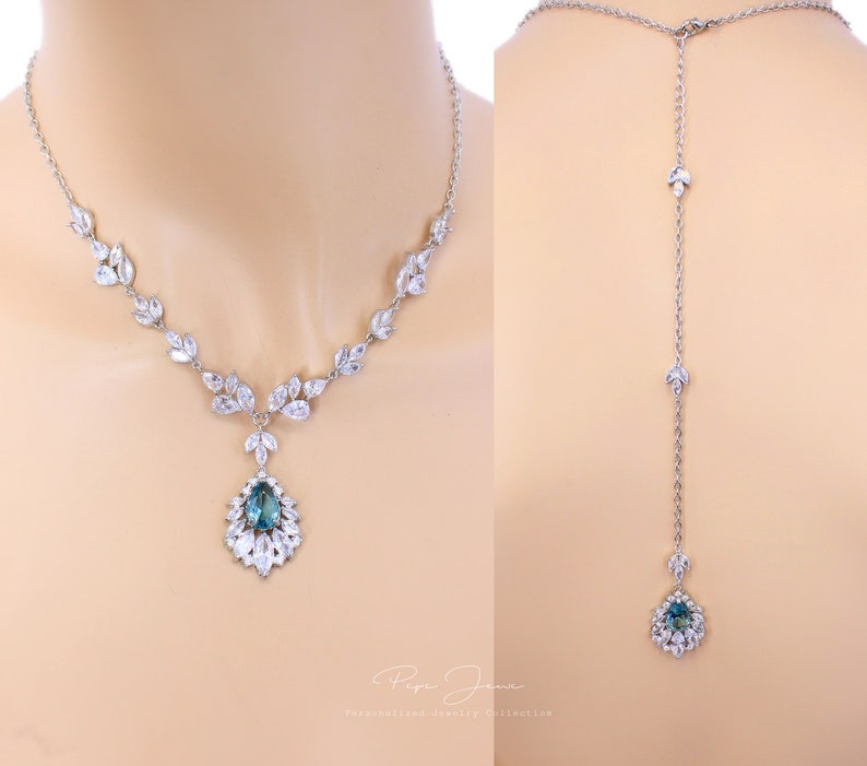 Puede incluir: Un collar de plata con un colgante de piedra preciosa azul en forma de l&aacute;grima rodeado de piedras transparentes m&aacute;s peque&ntilde;as. El collar tiene una cadena delicada con peque&ntilde;as piedras transparentes en forma de hojas. El collar se muestra en dos vistas: la delantera y la trasera. La vista trasera muestra el collar extendi&eacute;ndose hacia abajo por la espalda con una cadena m&aacute;s larga y una versi&oacute;n m&aacute;s peque&ntilde;a del colgante.