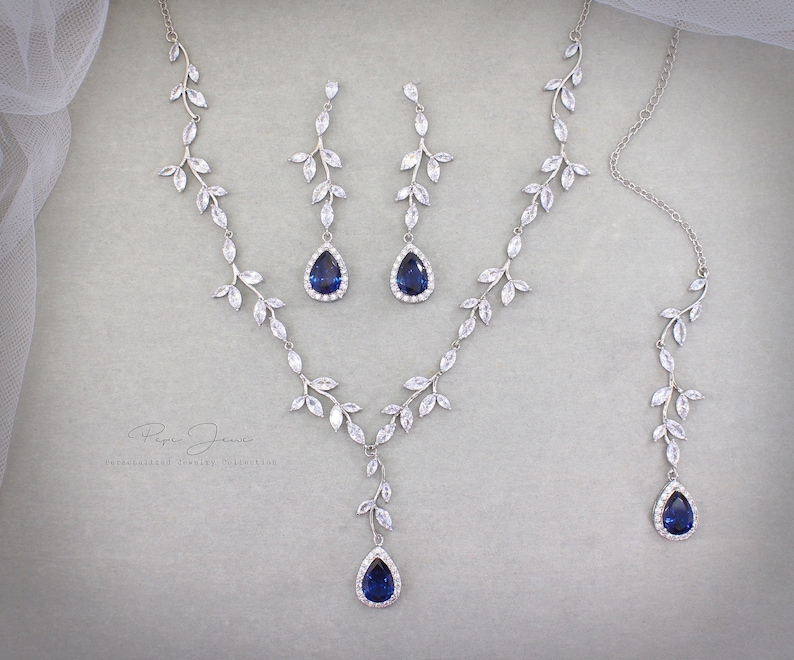May include: A silver necklace and earrings set with a blue teardrop-shaped stone and a leaf design. The jewelry is made of silver and cubic zirconia.