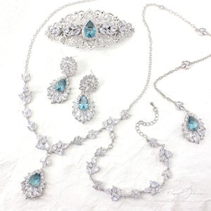 Puede incluir: Un conjunto de joyas de novia con una tiara plateada, un collar, una pulsera, pendientes y un peine para el cabello. El conjunto presenta piedras preciosas azules en forma de l&aacute;grima rodeadas de diamantes de imitaci&oacute;n transparentes.