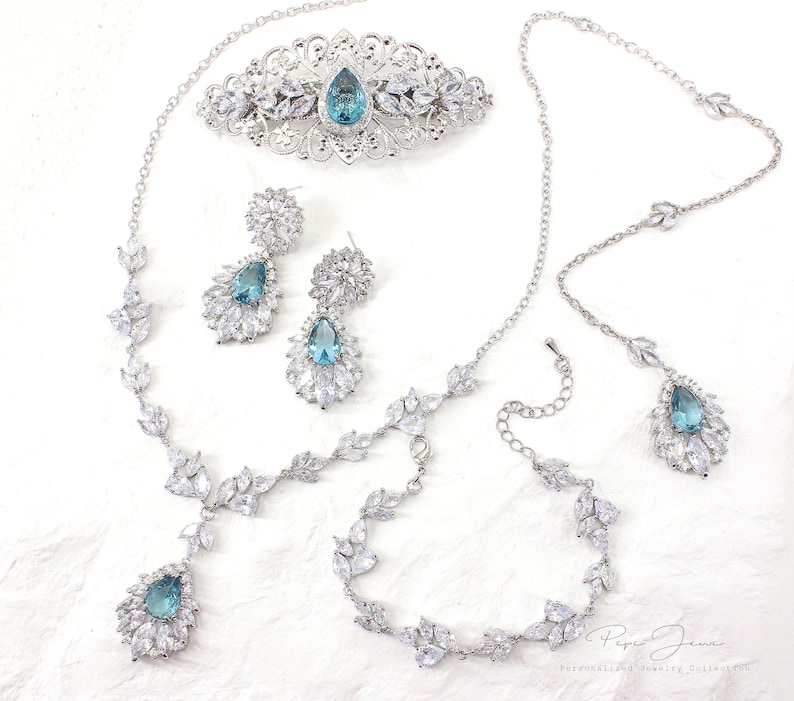 Puede incluir: Un conjunto de joyas de novia con una tiara plateada, un collar, una pulsera, pendientes y un peine para el cabello. El conjunto presenta piedras preciosas azules en forma de l&aacute;grima rodeadas de diamantes de imitaci&oacute;n transparentes.