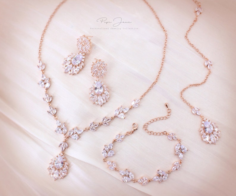 Puede incluir: Conjunto de joyas de novia de oro rosa con collar, pendientes y pulsera. Las joyas presentan un delicado dise&ntilde;o de hojas con piedras transparentes brillantes.