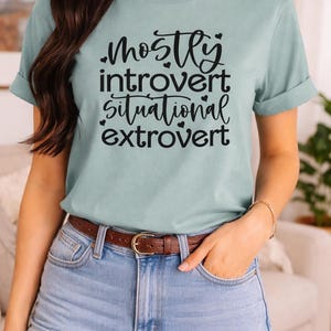May include: A light green t-shirt with the text "Mostly Introvert, Situational Extrovert" in black script. The shirt has rolled-up sleeves and is paired with denim shorts and a brown belt. A gold bracelet is visible on the wrist.