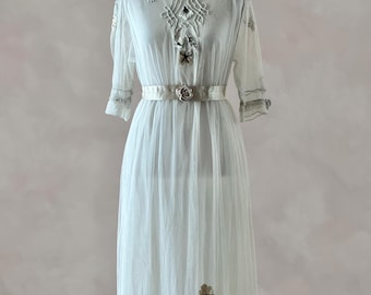 May include: A sheer white dress featuring elbow-length sleeves and a V-neckline. The bodice and hem are decorated with intricate silver and beige floral embroidery. A matching white satin ribbon belt with a central fabric rose cinches the waist.