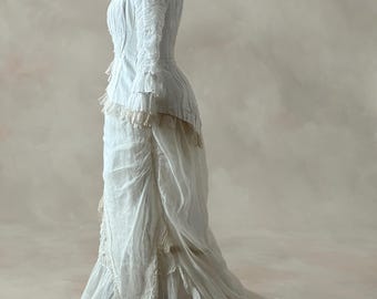 May include: A white vintage dress with a high collar, long sleeves, and a fitted bodice. The dress has detailed ruffles and lace trim. The skirt flows into a long train, creating an elegant silhouette. The dress is displayed on a mannequin.