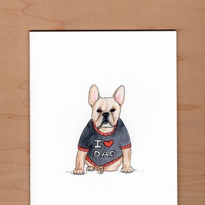 Pu&ograve; includere: Illustrazione ad acquerello di un bulldog francese con un maglione nero con finiture rosse. Il maglione ha la scritta "I ❤️ DAD" in lettere bianche con un cuore rosso. Il cane &egrave; marrone chiaro con il naso nero e si trova su uno sfondo bianco.