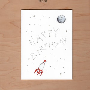 May include: A white card with a rocket ship, a moon, and stars. The words "Happy Birthday" are spelled out in a constellation of stars.